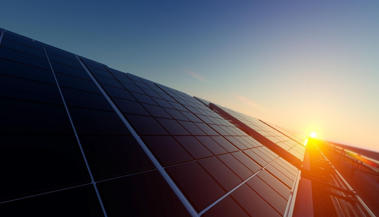 Overhead flat-lay photograph of dark solar panels angled toward a sunset sky with warm light flare, deep blue and amber tones.